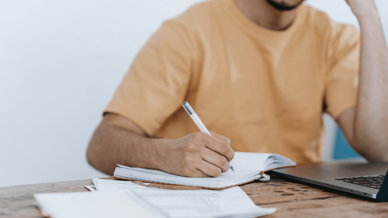Man with light orange shirt writing something on a piece of paper using his right hand